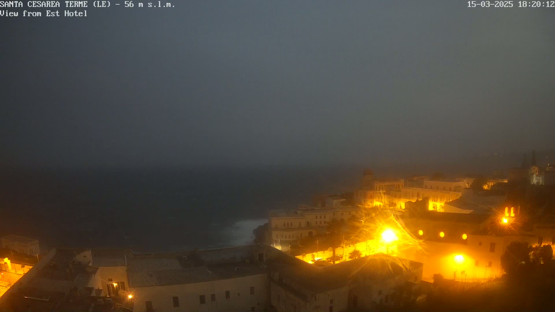 A coastal town is illuminated at night under a foggy, dark sky, with waves visible in the dark ocean below.