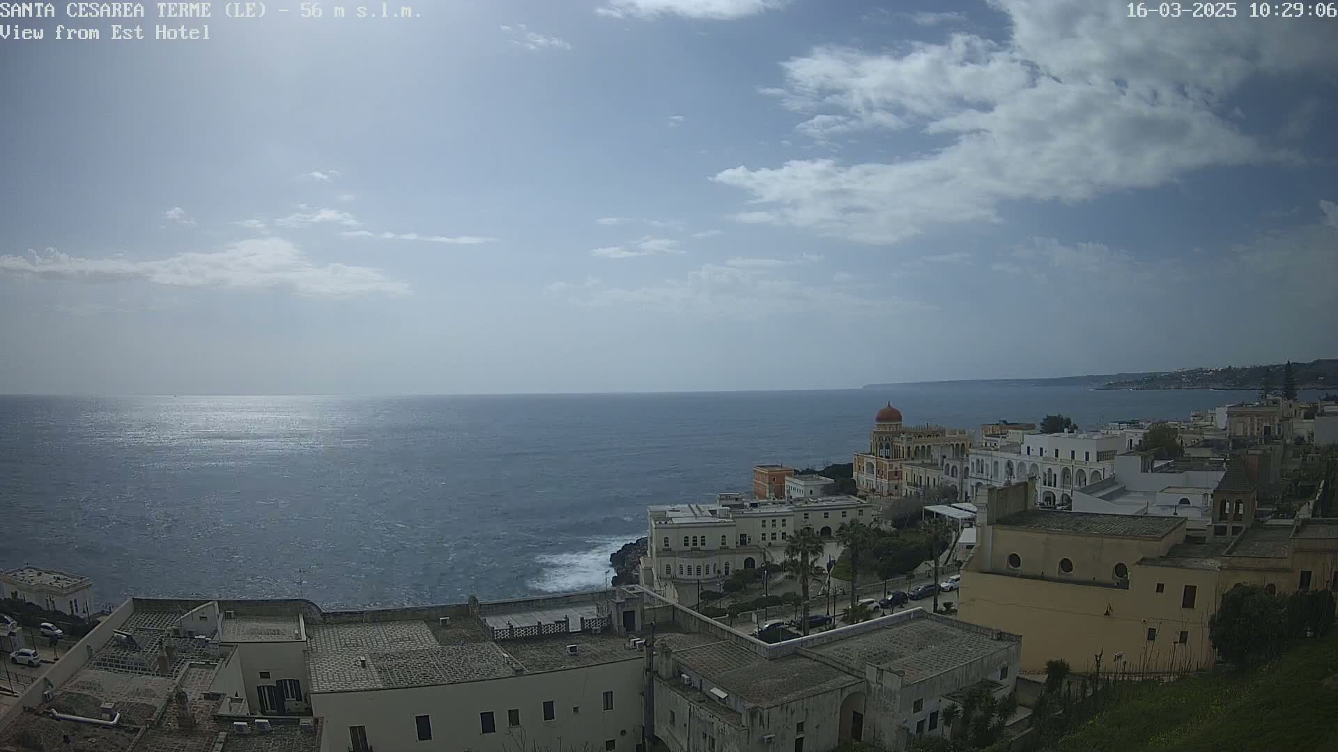 A partly cloudy sky overlooks a coastal town with white buildings and a calm, blue sea.