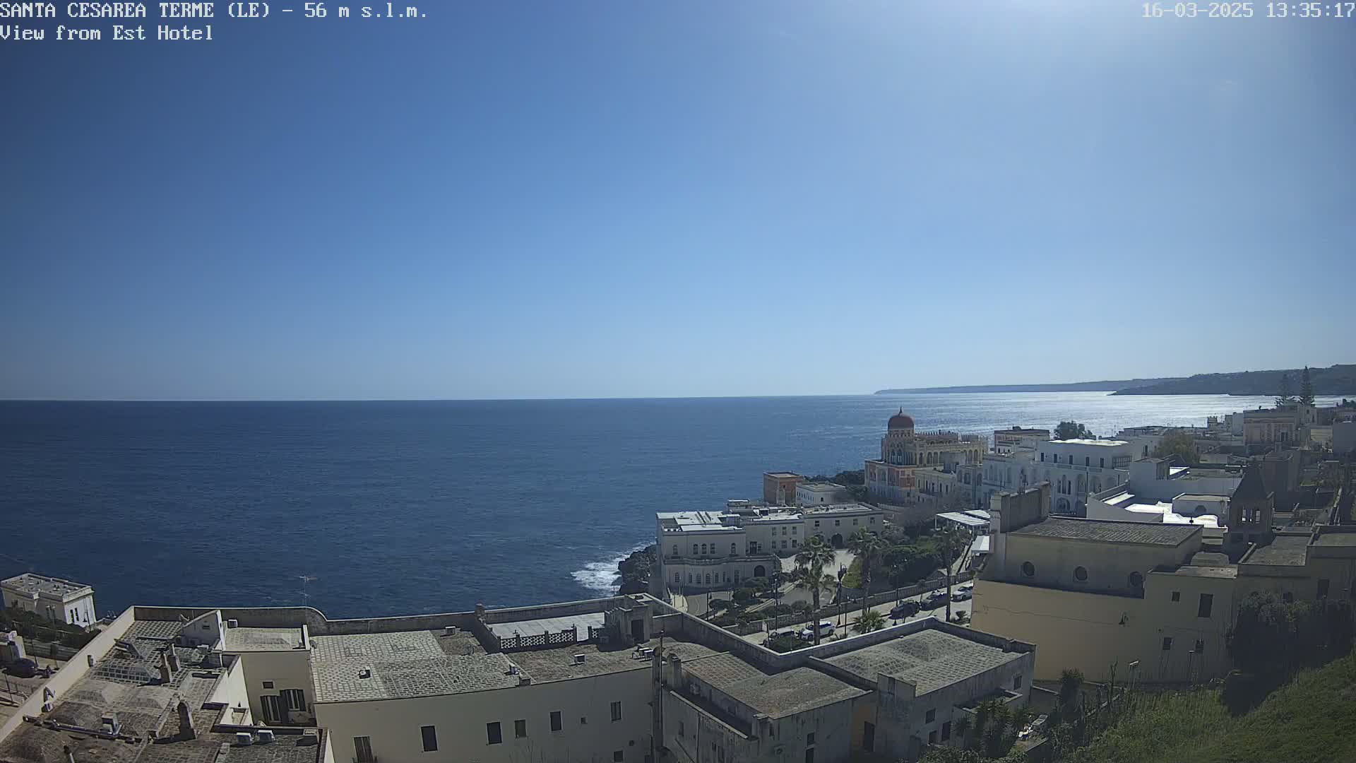 A coastal town with white buildings, a church dome, and palm trees sits on a cliff overlooking a calm, blue sea under a clear, sunny sky.