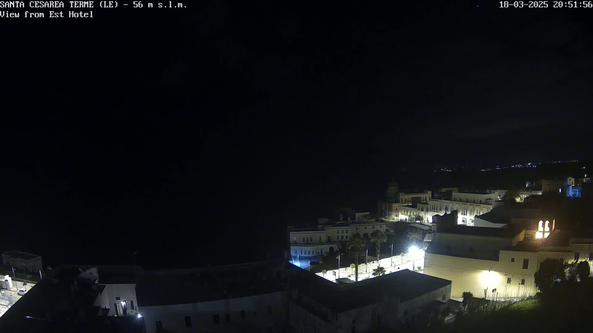 A nighttime view of a coastal town with buildings lit up, overlooking a dark body of water under a clear, dark sky.