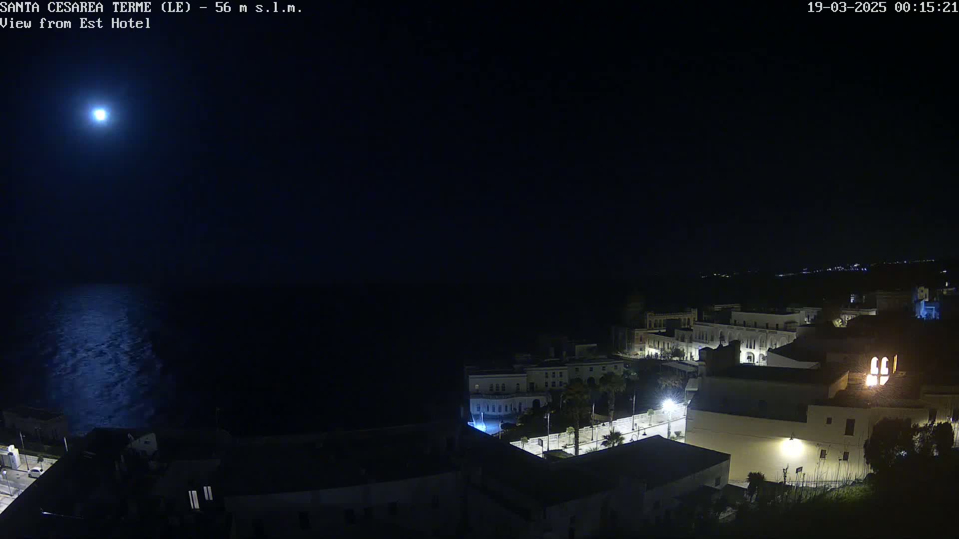 A bright moon illuminates a dark seaside town at night, its calm waters reflecting the lunar glow.