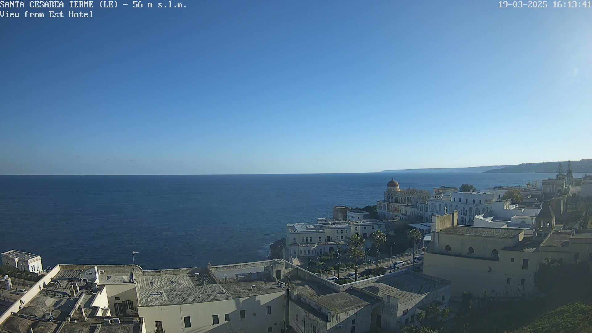 A coastal town with white buildings sits on a cliff overlooking a calm blue sea under a clear blue sky.