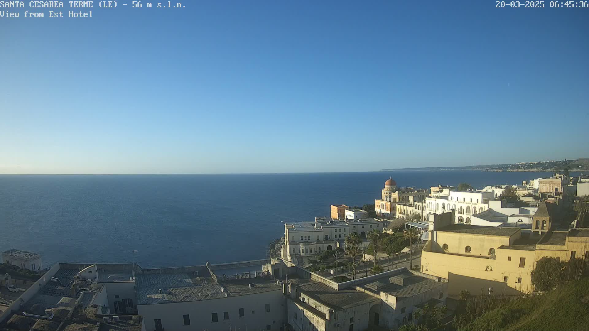 A coastal town with white buildings and a domed structure overlooks a calm, blue sea under a clear blue sky.
