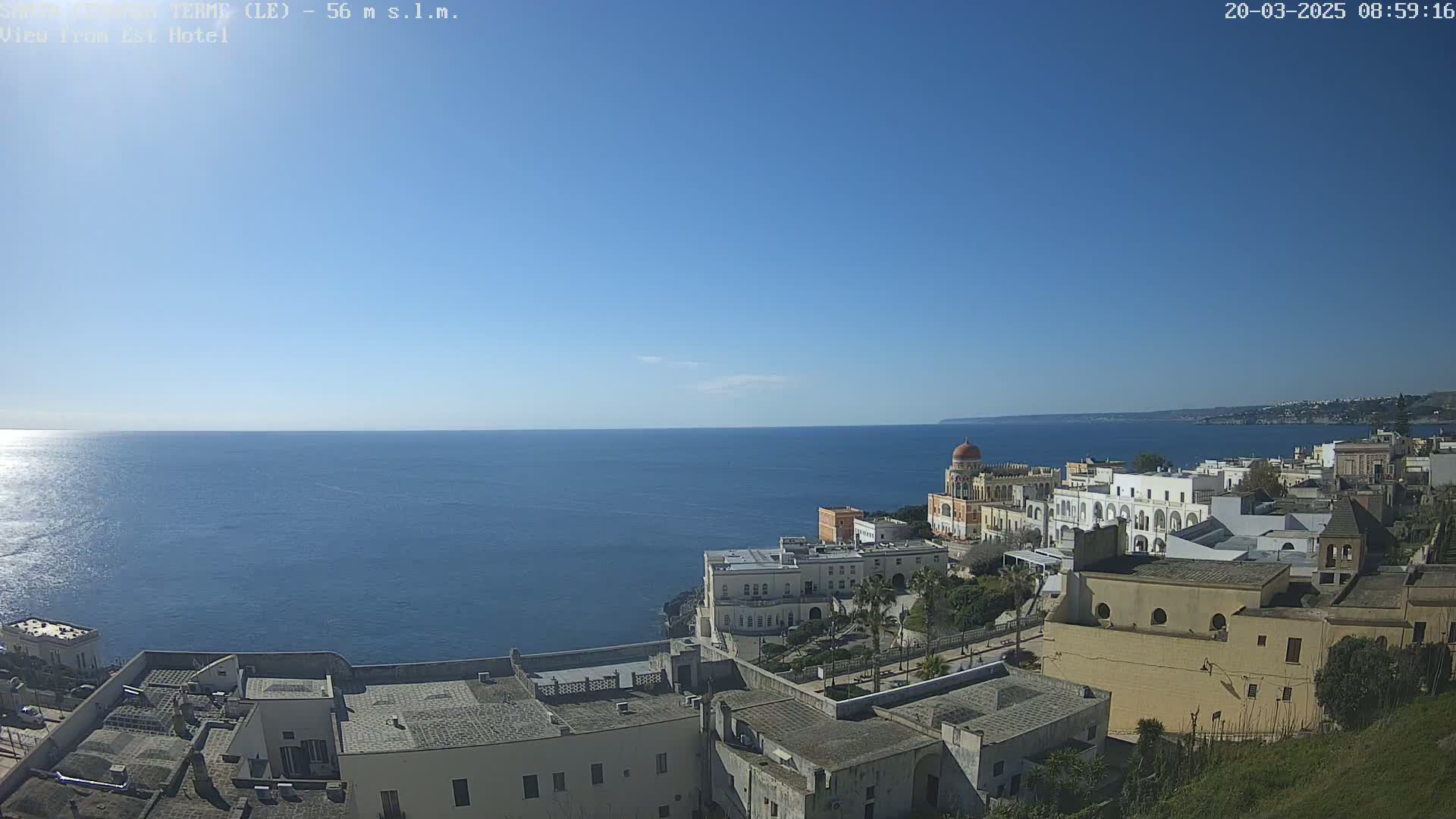 A coastal town with white buildings and a calm blue sea under a clear blue sky is viewed from a high vantage point.