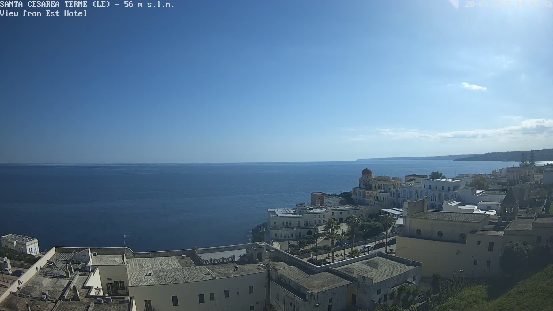 A coastal town with white buildings overlooks a calm, blue sea under a mostly clear, sunny sky.