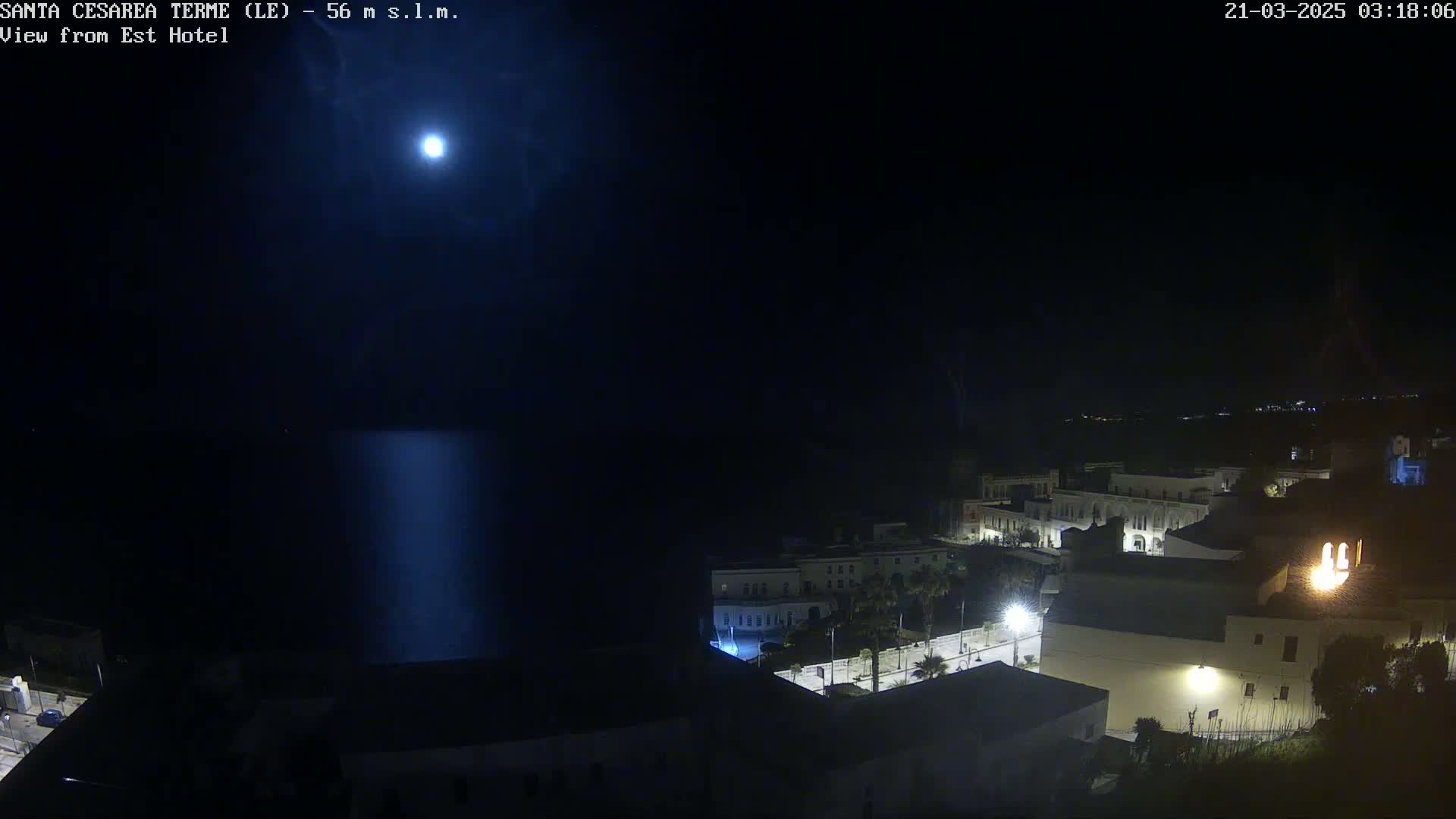 A nighttime, moonlit view of a coastal town with a calm sea reflecting light.