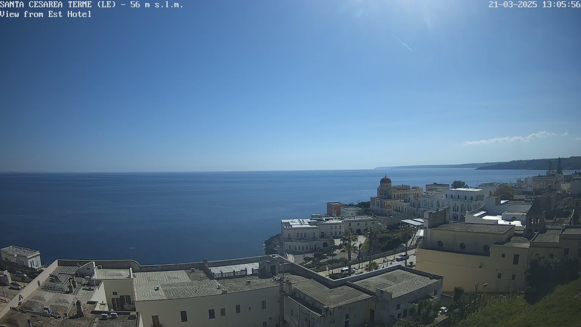 A coastal town with white buildings overlooks a calm, blue sea under a clear blue sky with a few wispy clouds.