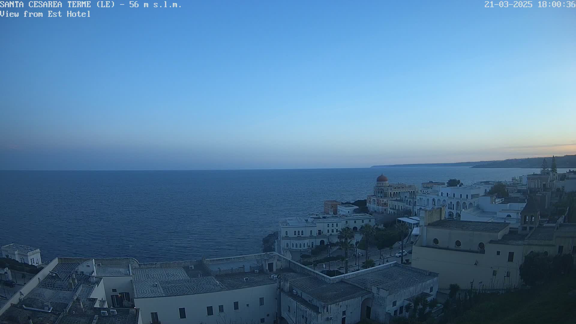 A coastal town with white buildings, a domed structure, and palm trees overlooks a calm, dark blue sea under a clear twilight sky.