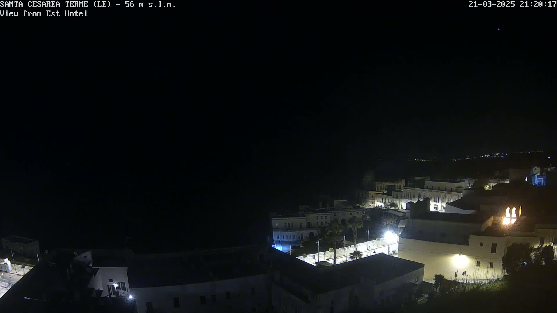 A nighttime aerial view of a coastal town with dimly lit buildings, streets, and palm trees, under a clear, dark sky.