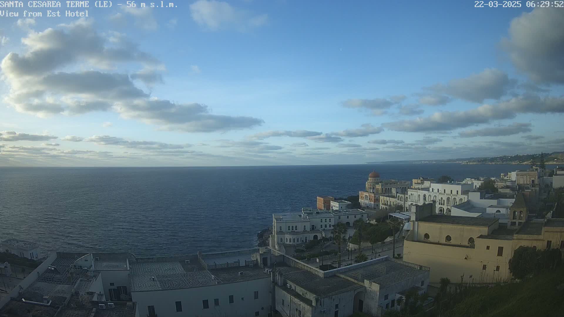 A coastal town with white buildings overlooks a calm sea under a partly cloudy sky.