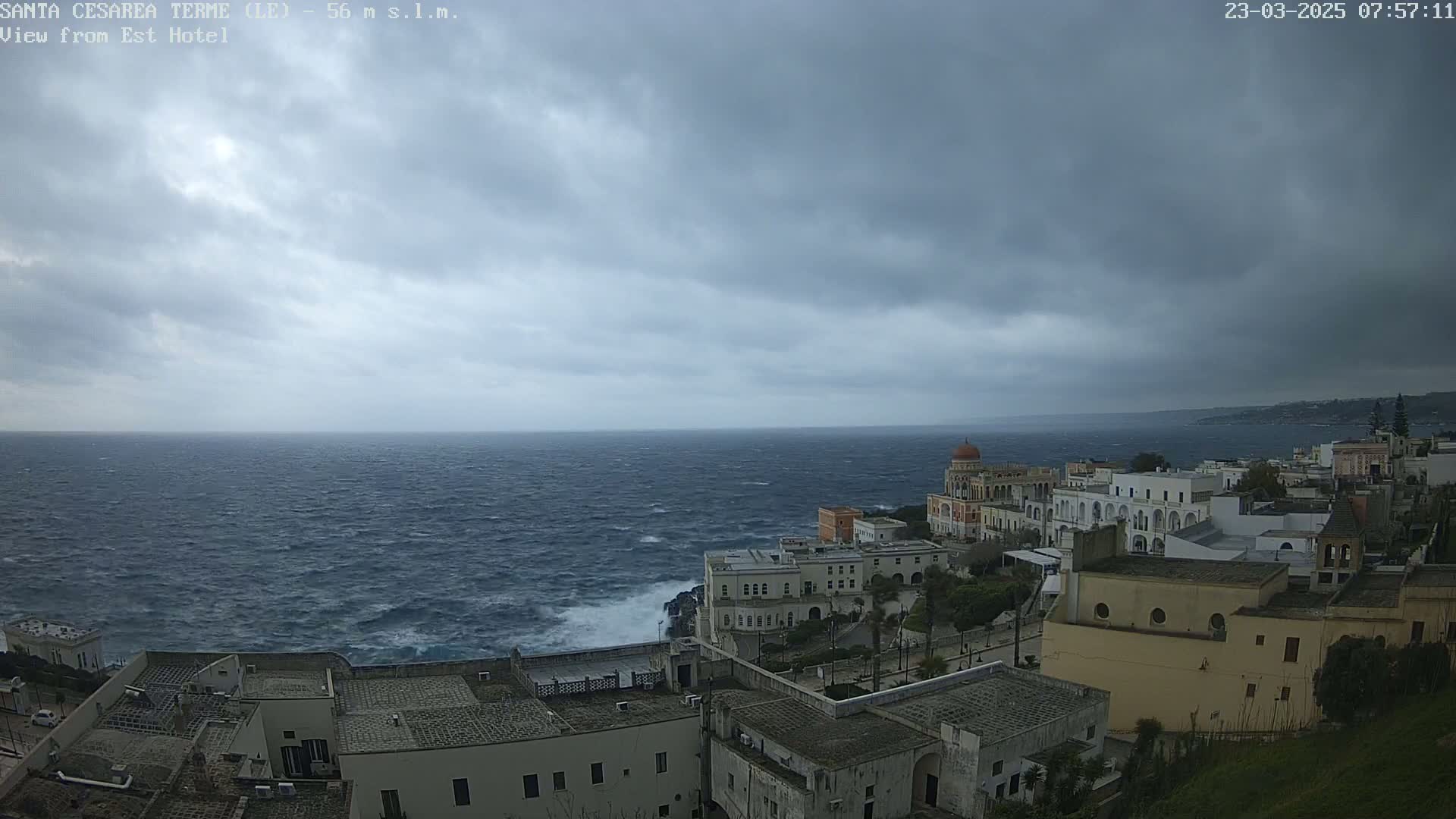 A coastal town with white buildings overlooks a rough, dark-blue sea under a cloudy, overcast sky.