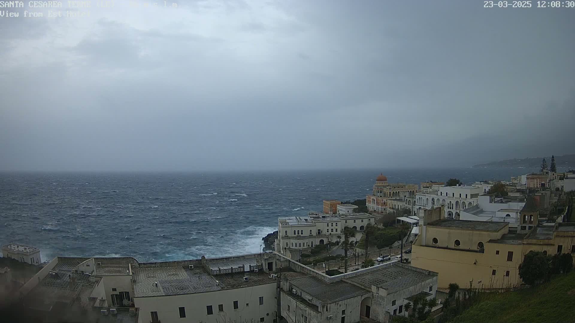 A coastal town with white buildings is shown under an overcast sky, with a rough, choppy sea visible in the background.
