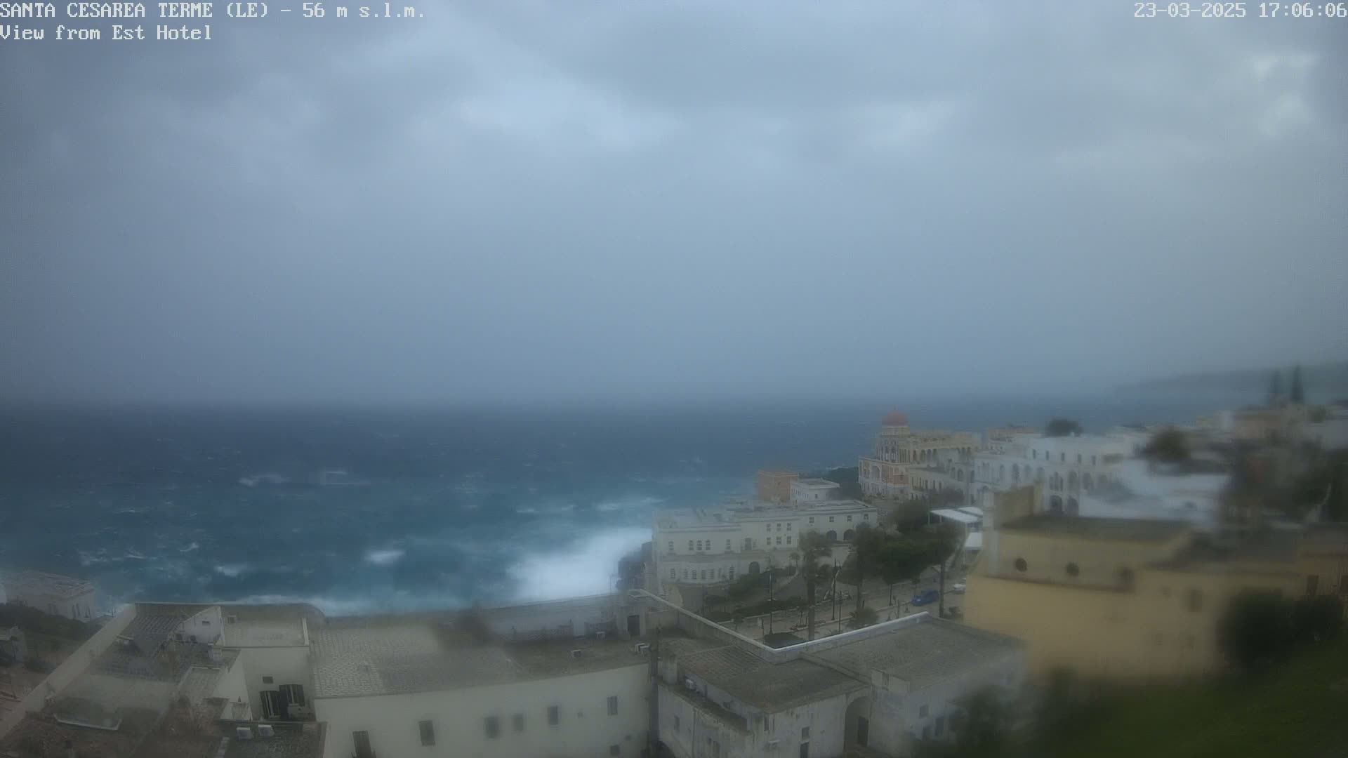 A coastal town with white buildings is shown under an overcast sky, with a rough sea visible in the background.