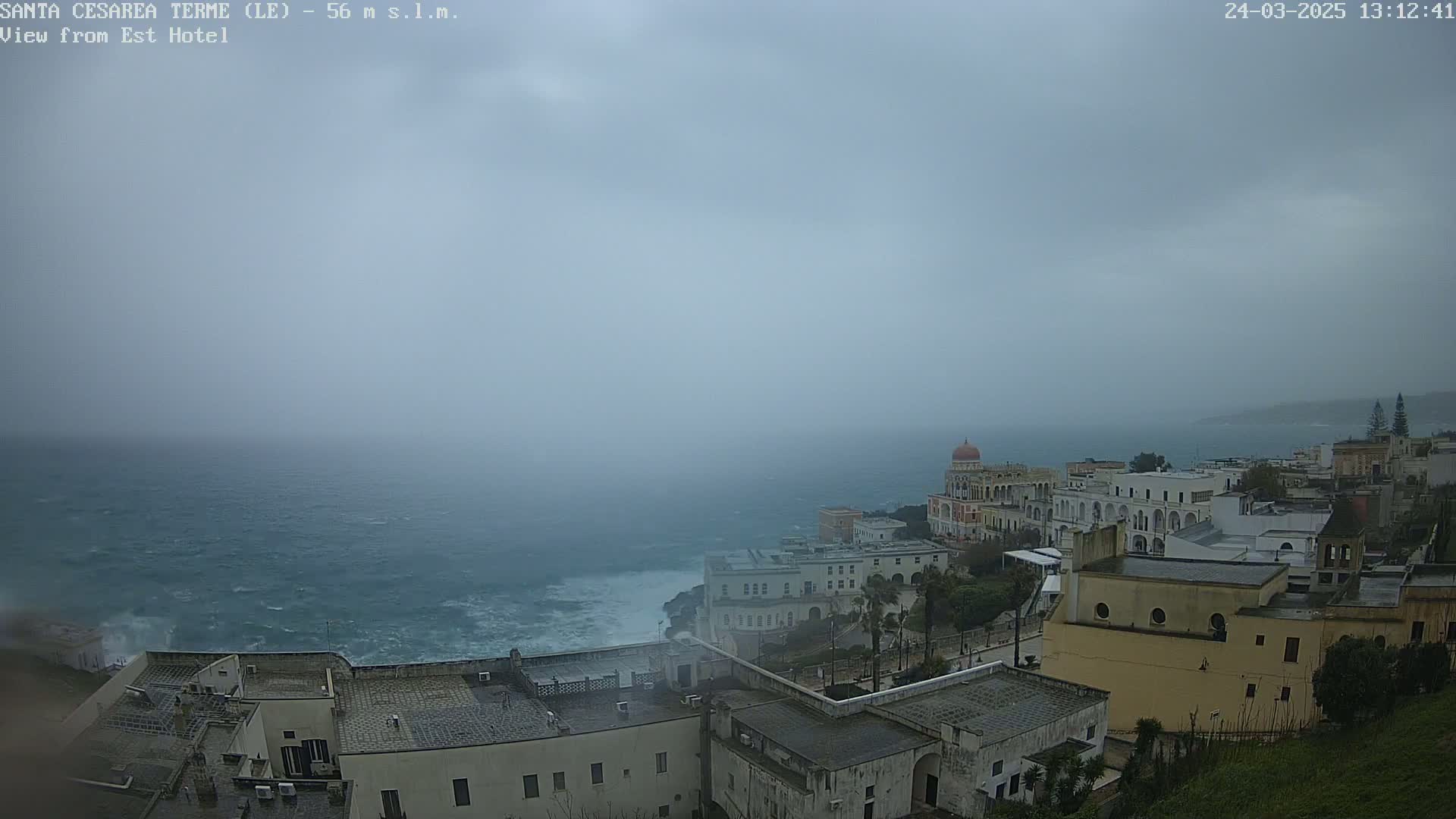 A coastal town with white buildings overlooks a rough sea under an overcast sky.