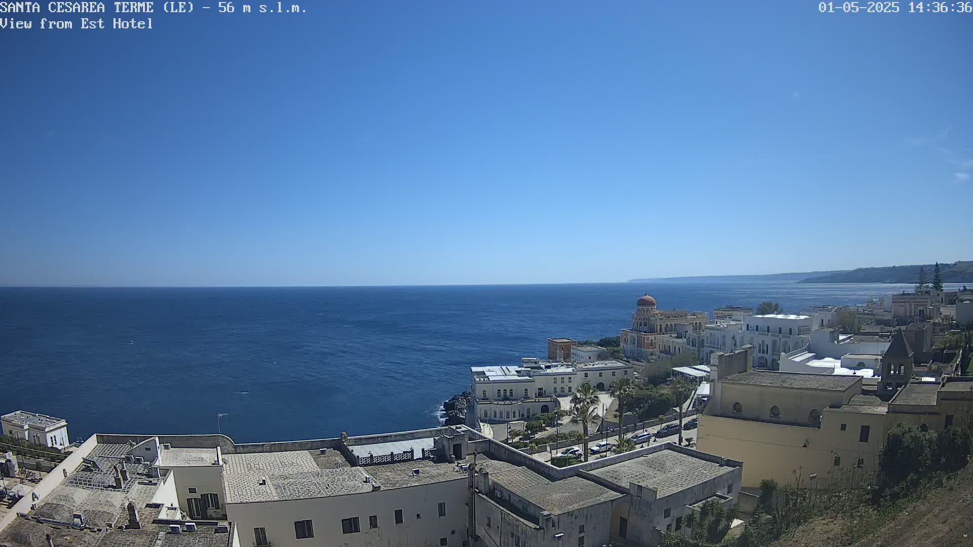 A coastal town with white buildings and a domed structure overlooks a calm, blue sea under a clear, sunny sky.