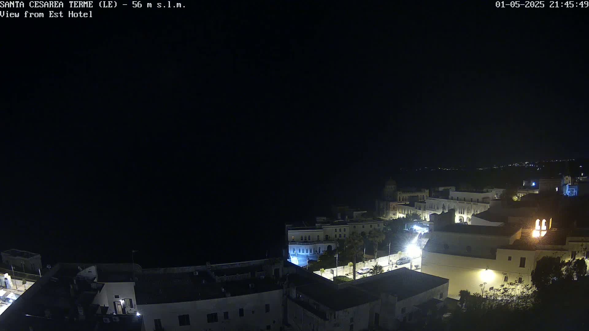 A nighttime, high-angle view of a coastal town with sparsely lit buildings and streets, overlooking a dark body of water under a clear, dark sky.