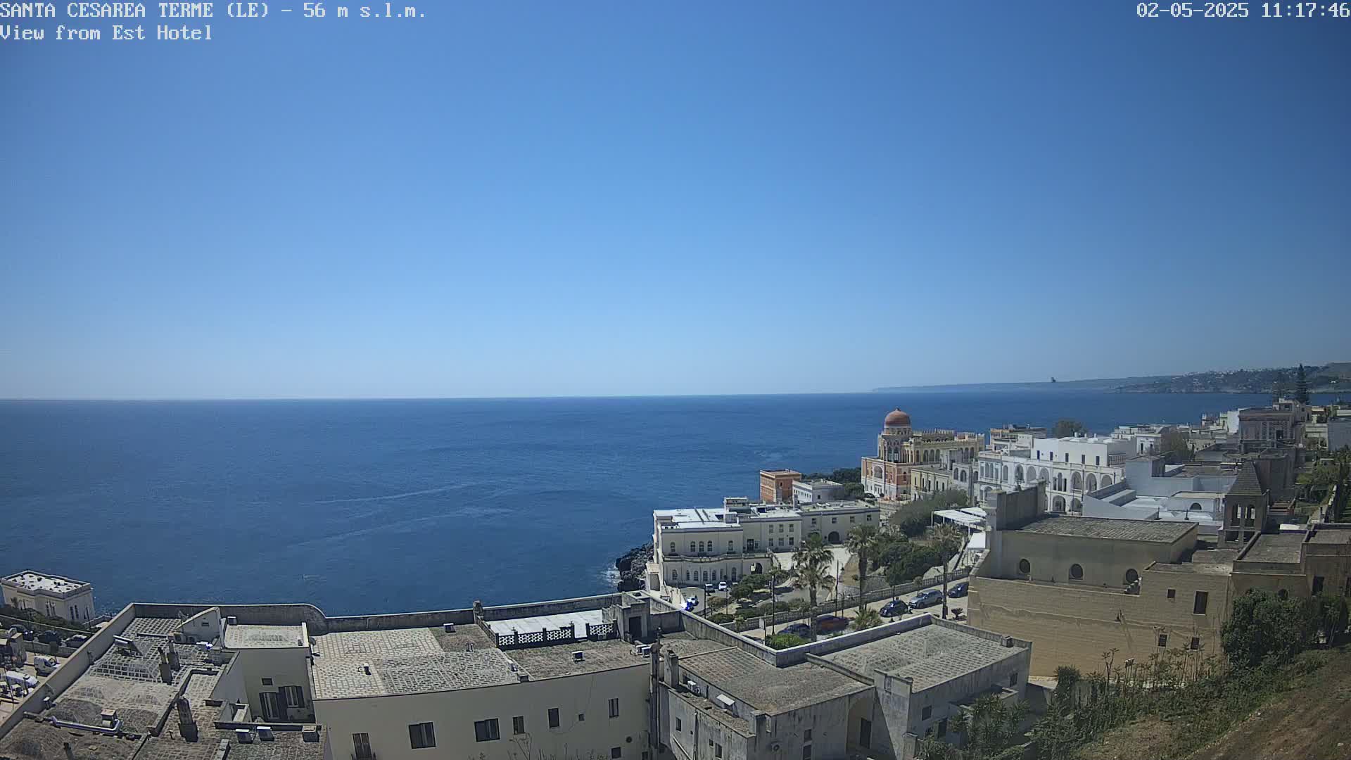 A coastal town with white buildings and a domed structure overlooks a calm blue sea under a clear blue sky.