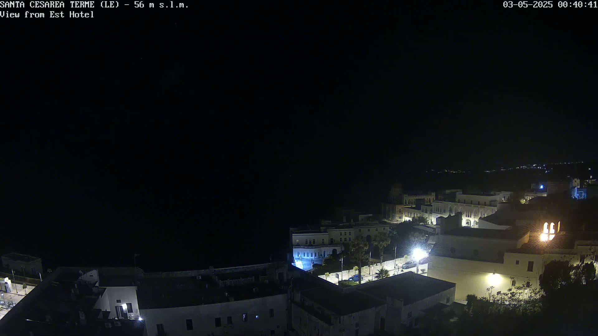 A nighttime, high-angle view shows a coastal town with dimly lit buildings and a dark sea in the background.
