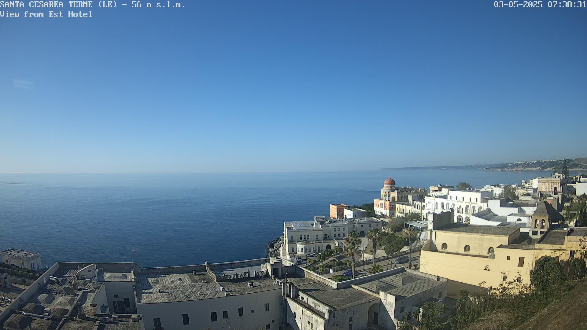 A coastal town with white buildings and a domed structure overlooks a calm, blue sea under a clear, bright blue sky.