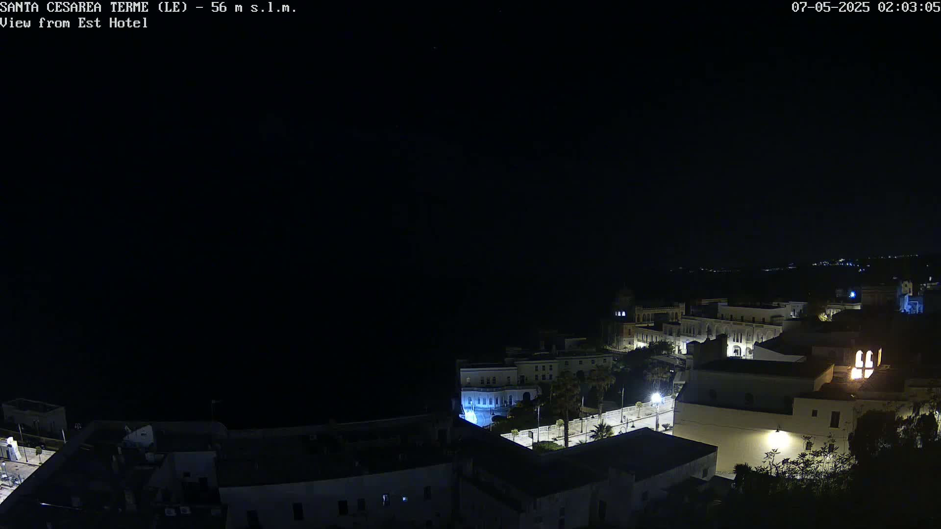 A nighttime, high-angle view of a coastal town with dimly lit buildings and streets near the dark sea under a clear, starlit sky.