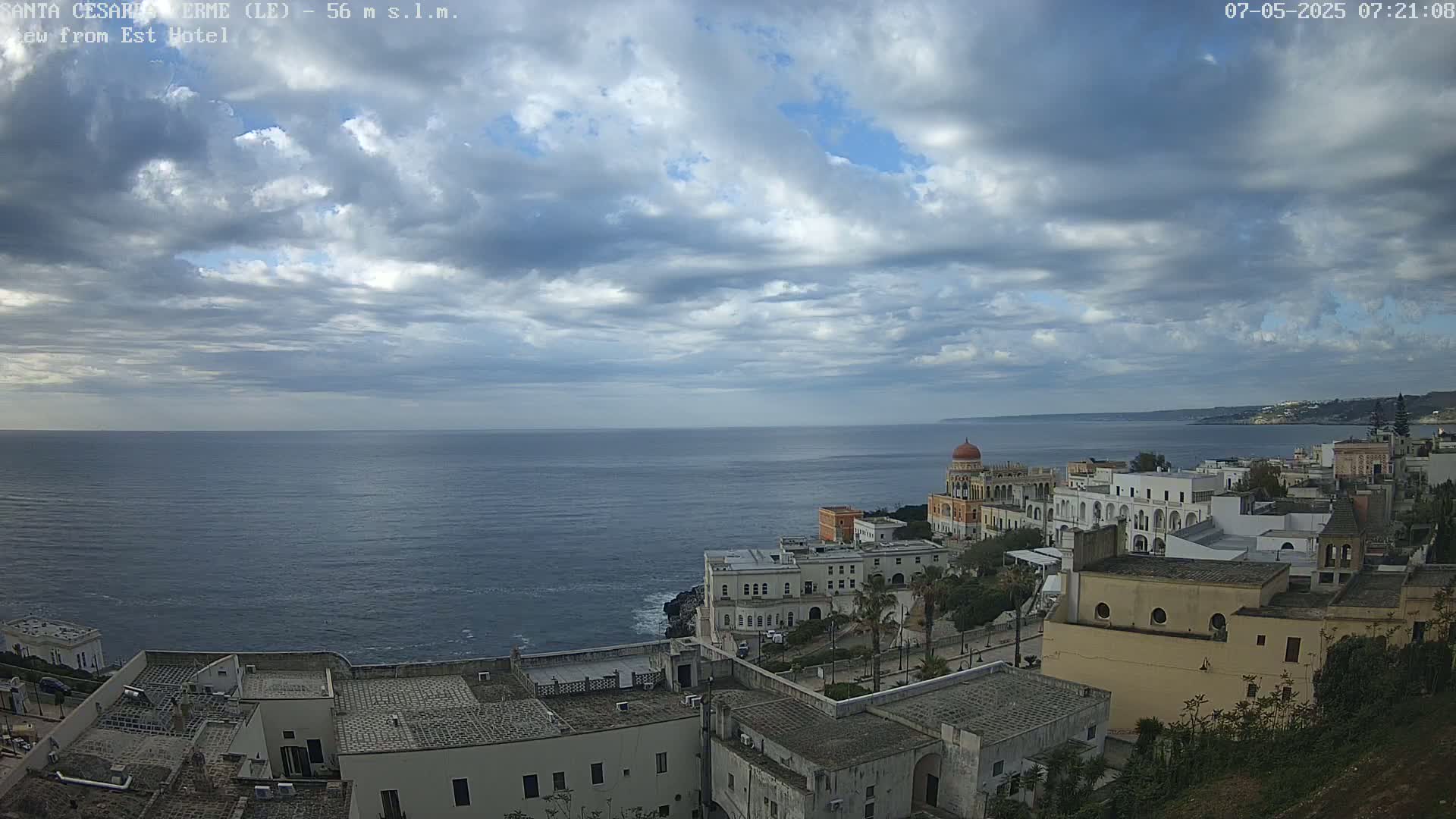 A coastal town with white buildings sits on a cliff overlooking a calm sea under a partly cloudy sky.