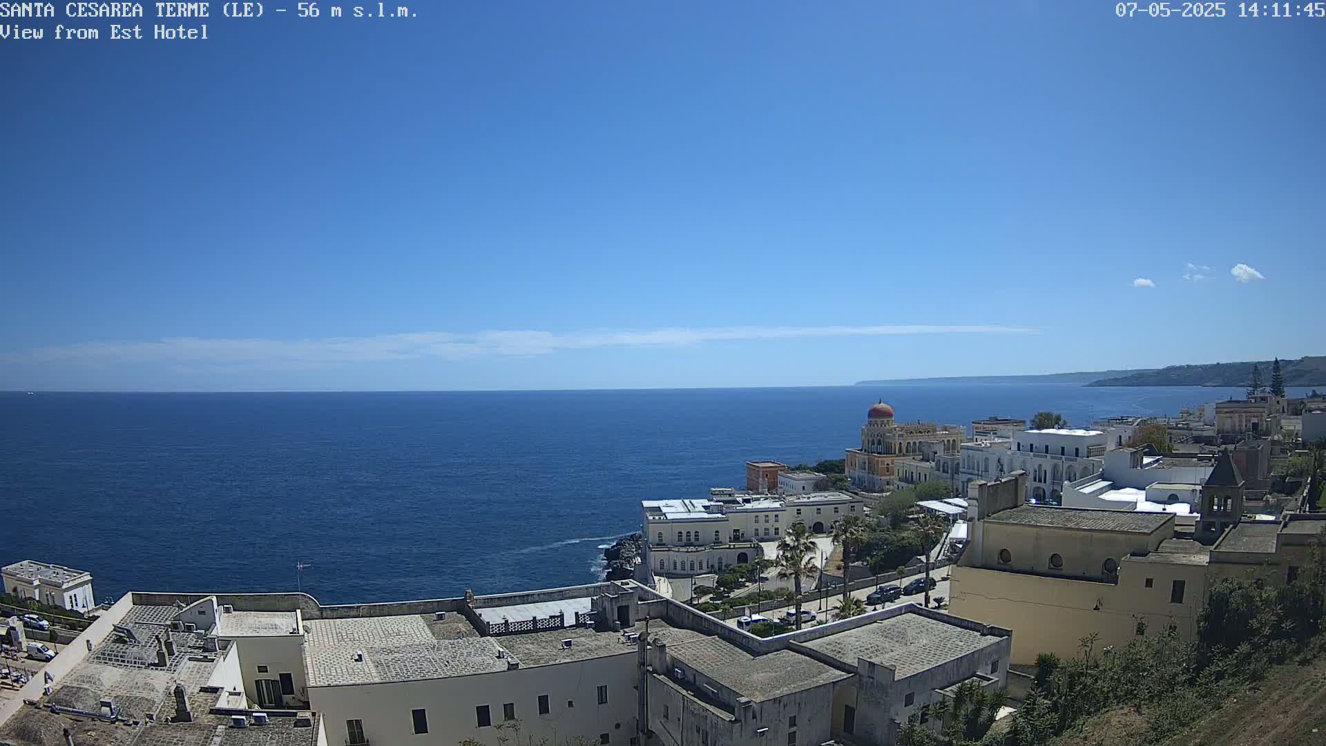 A coastal town with white buildings and a red-domed structure overlooks a calm blue sea under a clear blue sky.