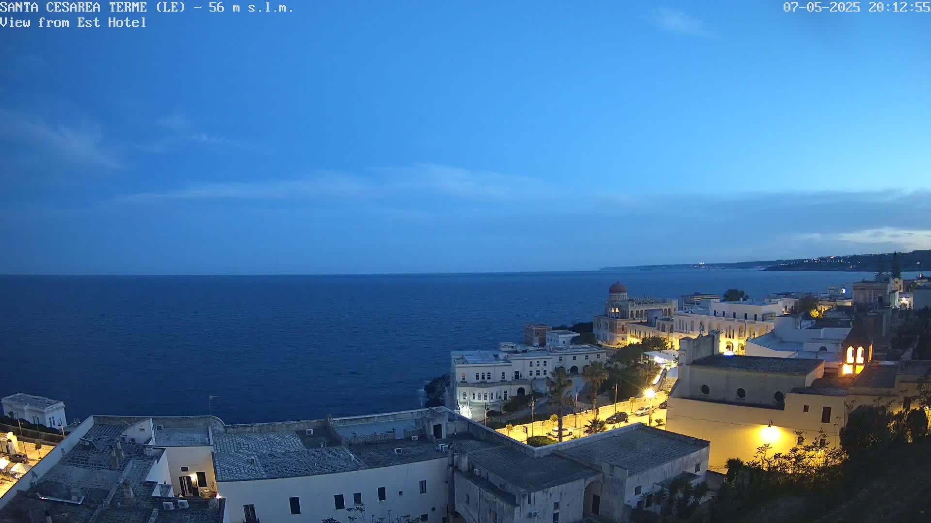 A coastal town at twilight is illuminated against a dark blue sea under a mostly clear sky.