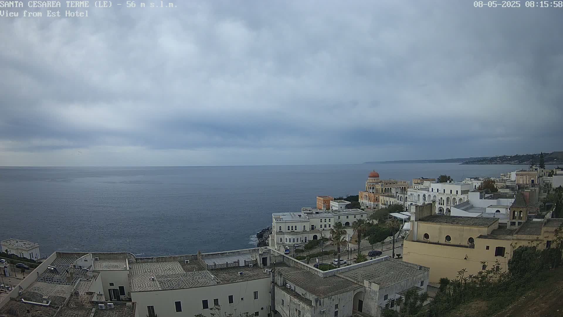 A coastal town with white buildings and a domed structure overlooks a calm, gray sea under an overcast sky.