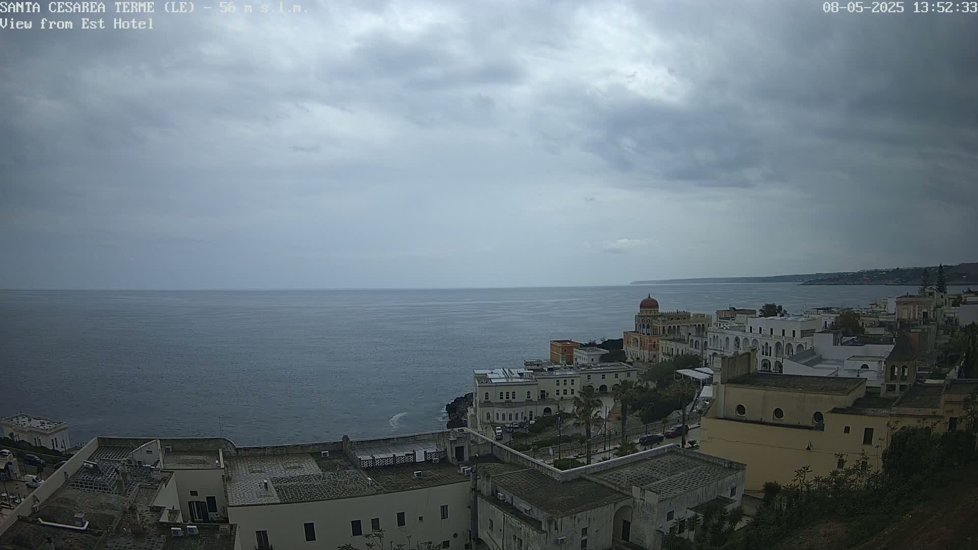 A coastal town with white buildings under a cloudy sky overlooks a calm sea.