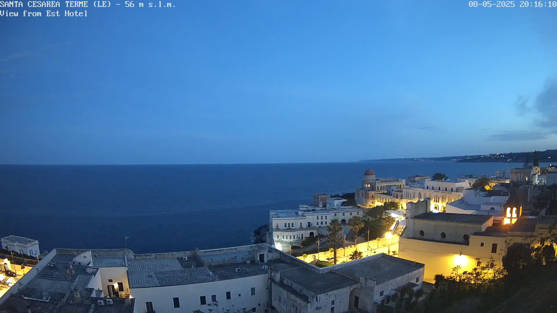 A coastal town at twilight is illuminated, overlooking a dark blue sea under a clear, dusk sky.