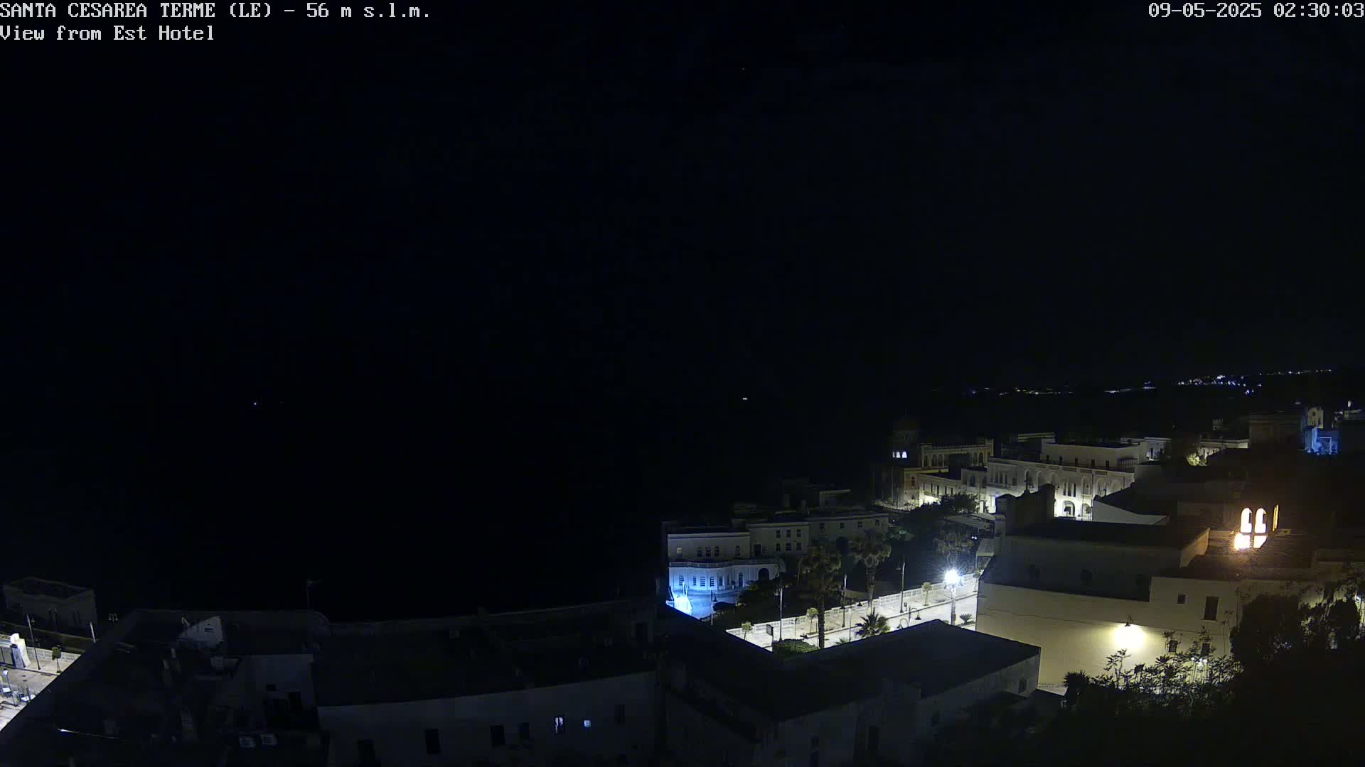 A nighttime, high-angle view of a coastal town with dimly lit buildings and streets, overlooking a dark body of water under a clear, dark sky.