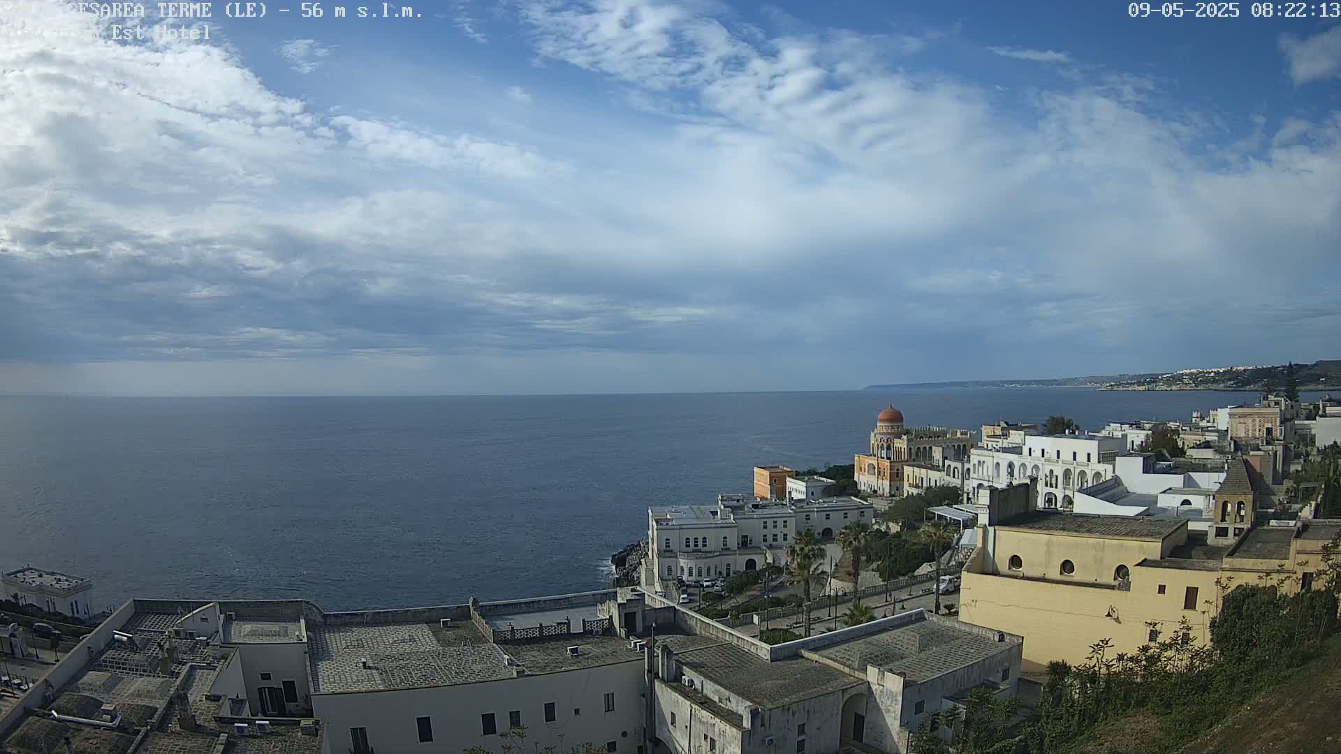 A coastal town with white buildings and a domed structure overlooks a calm sea under a partly cloudy sky.