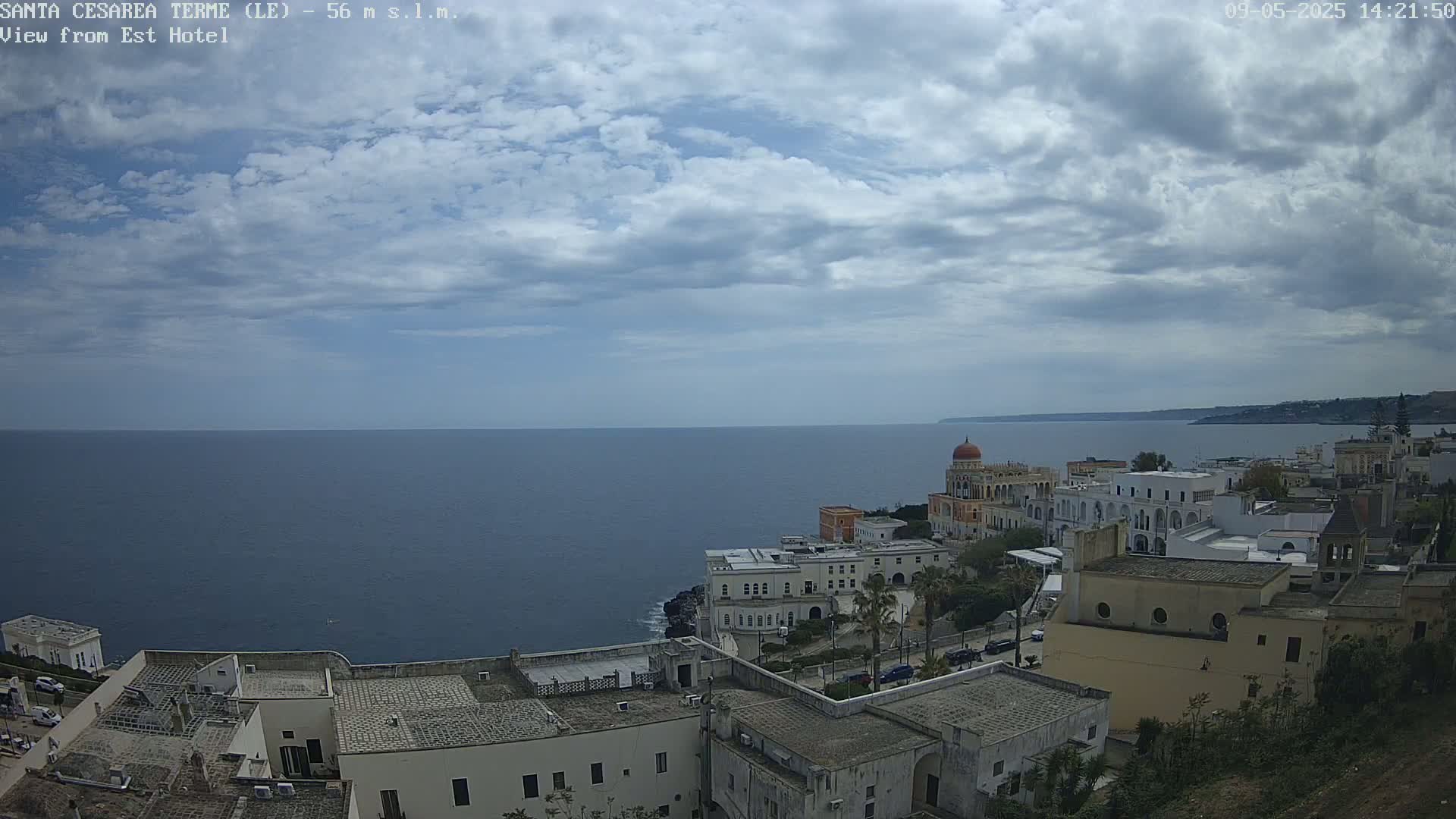 A coastal town with white buildings, a prominent domed structure, and a calm sea under a partly cloudy sky.