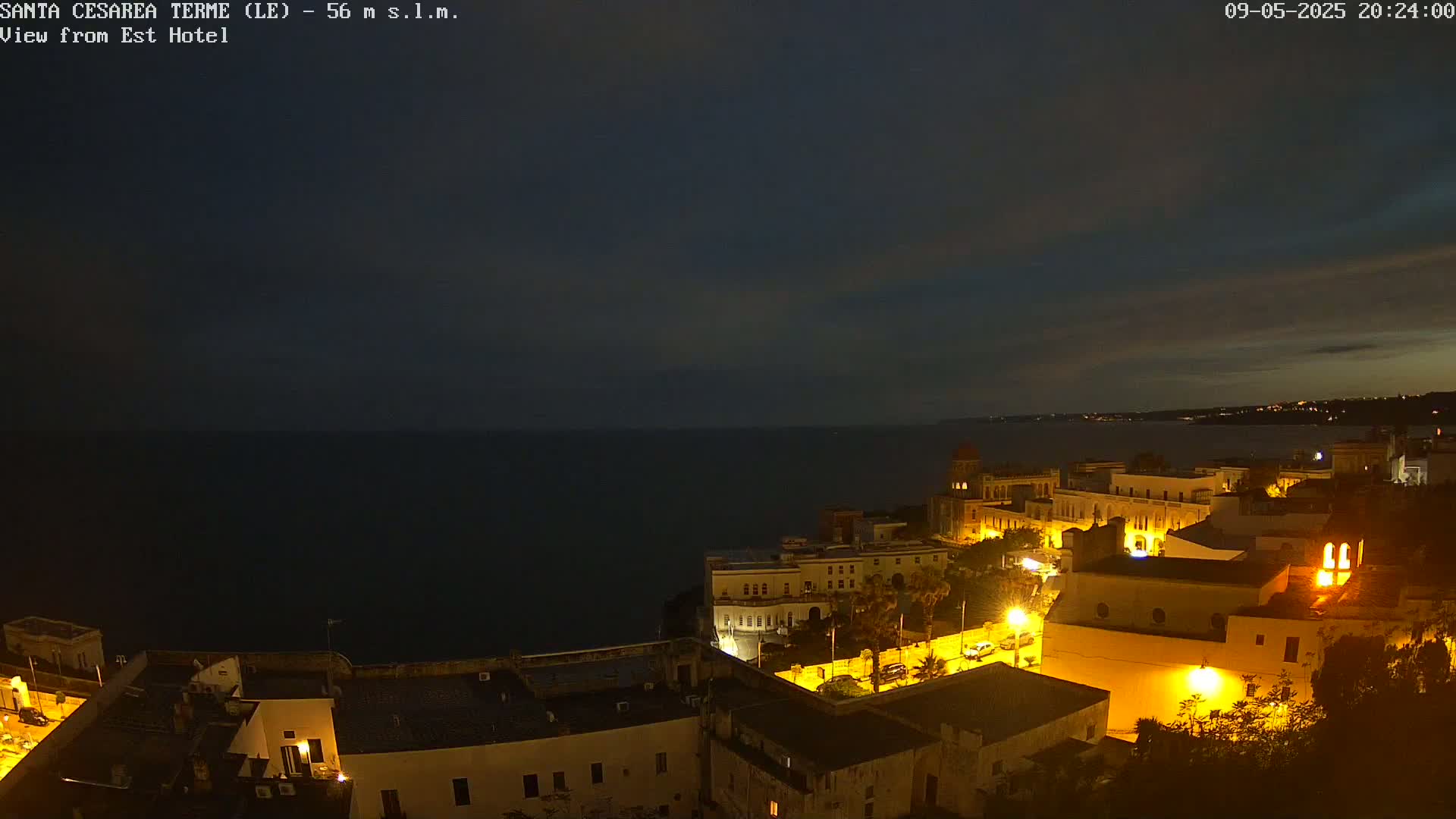 A nighttime coastal town is illuminated, overlooking a dark sea under a mostly clear, dark sky.