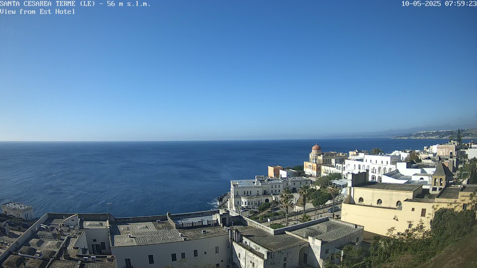 A coastal town with white buildings and a domed structure overlooks a calm, blue sea under a clear, bright blue sky.