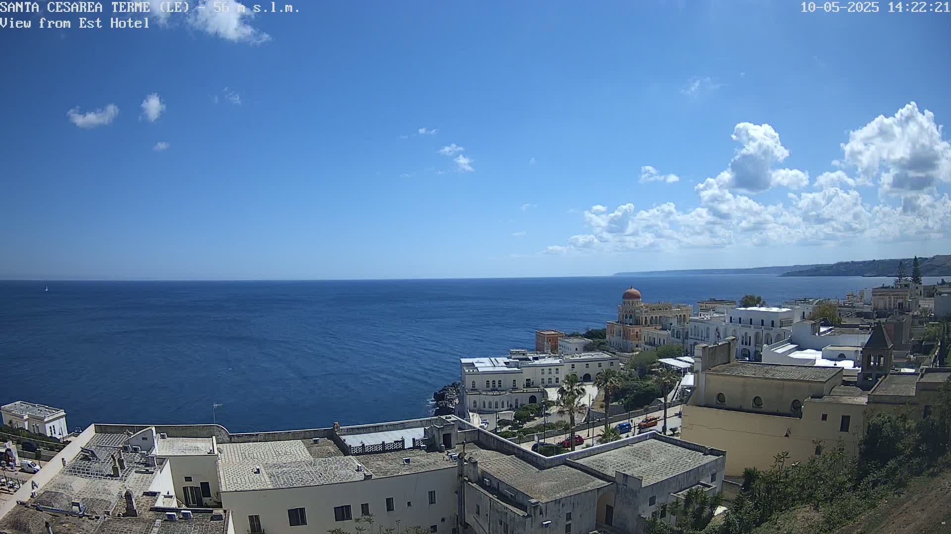A coastal town with white buildings sits on a cliff overlooking a calm blue sea under a mostly sunny sky with scattered cumulus clouds.