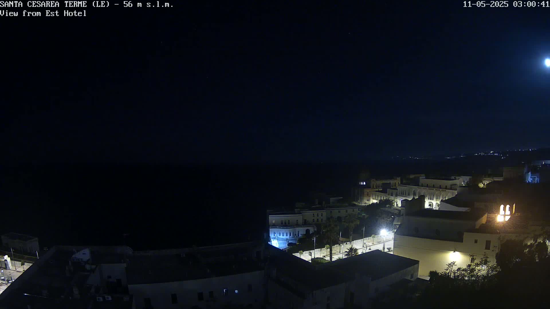 A nighttime, coastal town scene is illuminated under a bright moon, showing a dark sea and buildings with some lights.