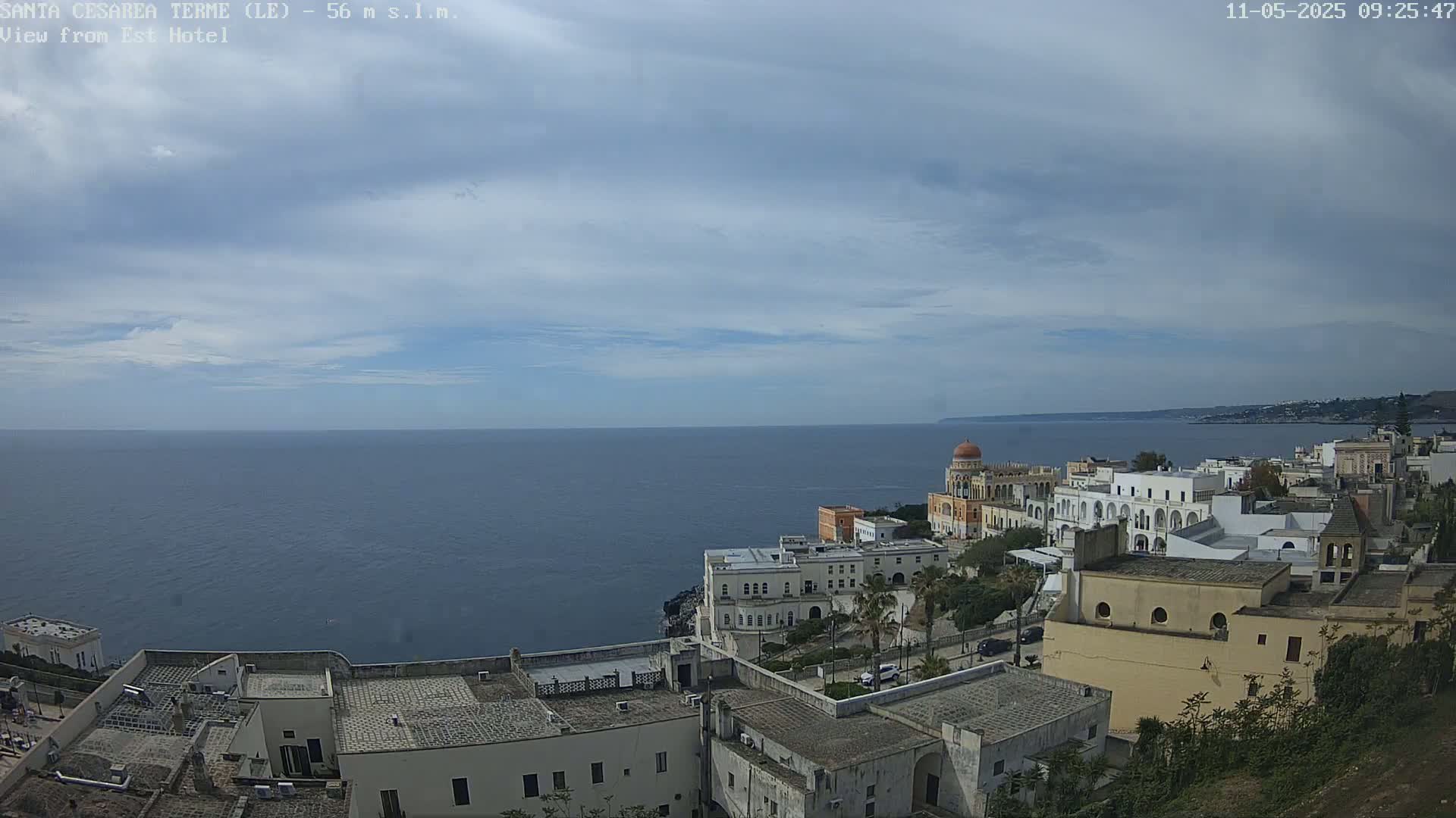 A coastal town with white buildings sits on a cliff overlooking a calm sea under a mostly cloudy sky.