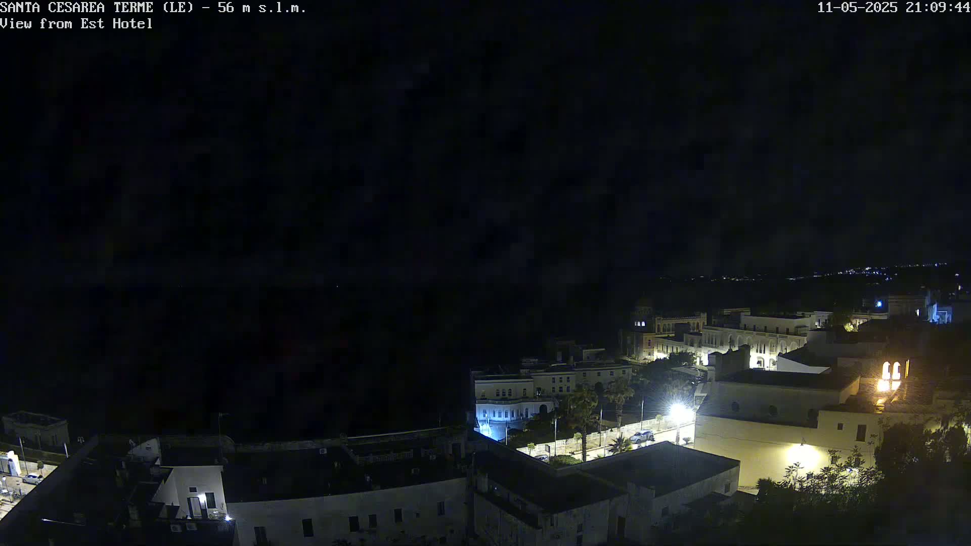 A nighttime, high-angle view of a coastal town with dimly lit buildings and a dark sea in the background.