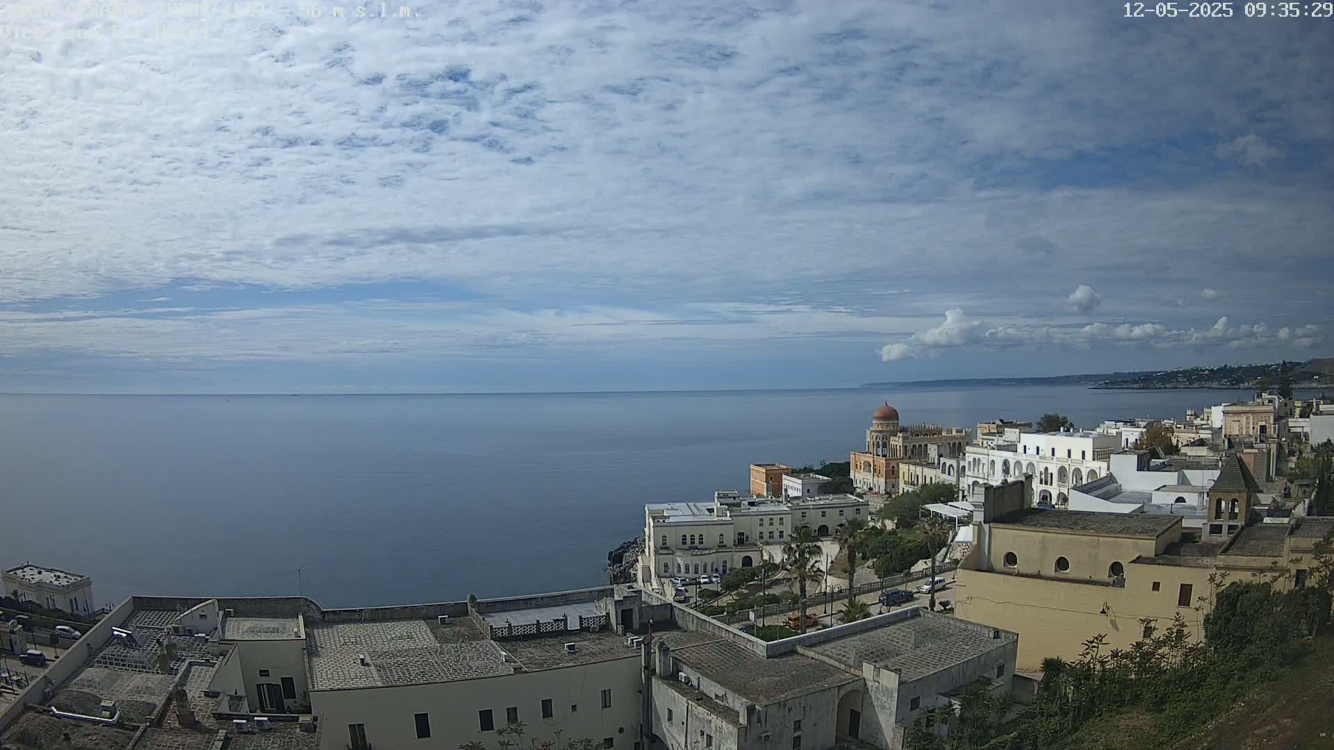 A coastal town with white buildings and a domed structure overlooks a calm sea under a partly cloudy sky.