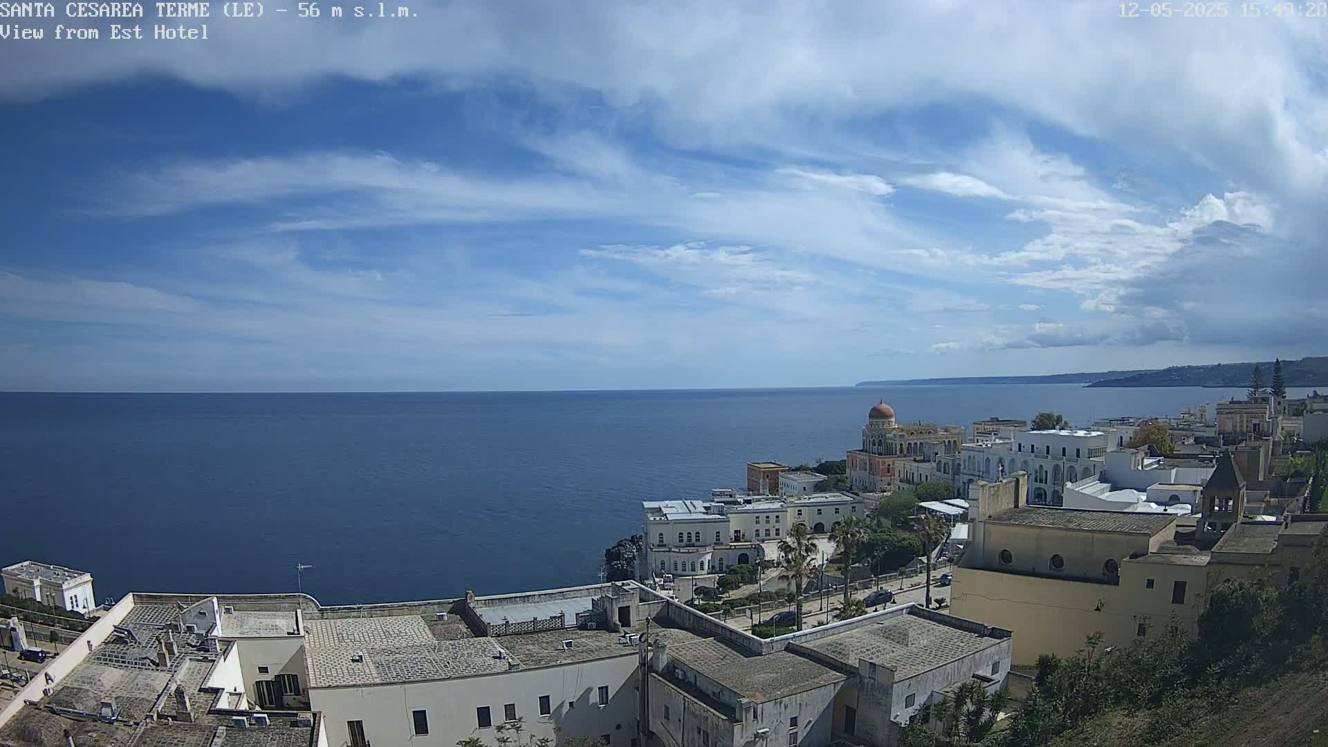 A coastal town with white buildings and a domed structure overlooks a calm blue sea under a partly cloudy sky.