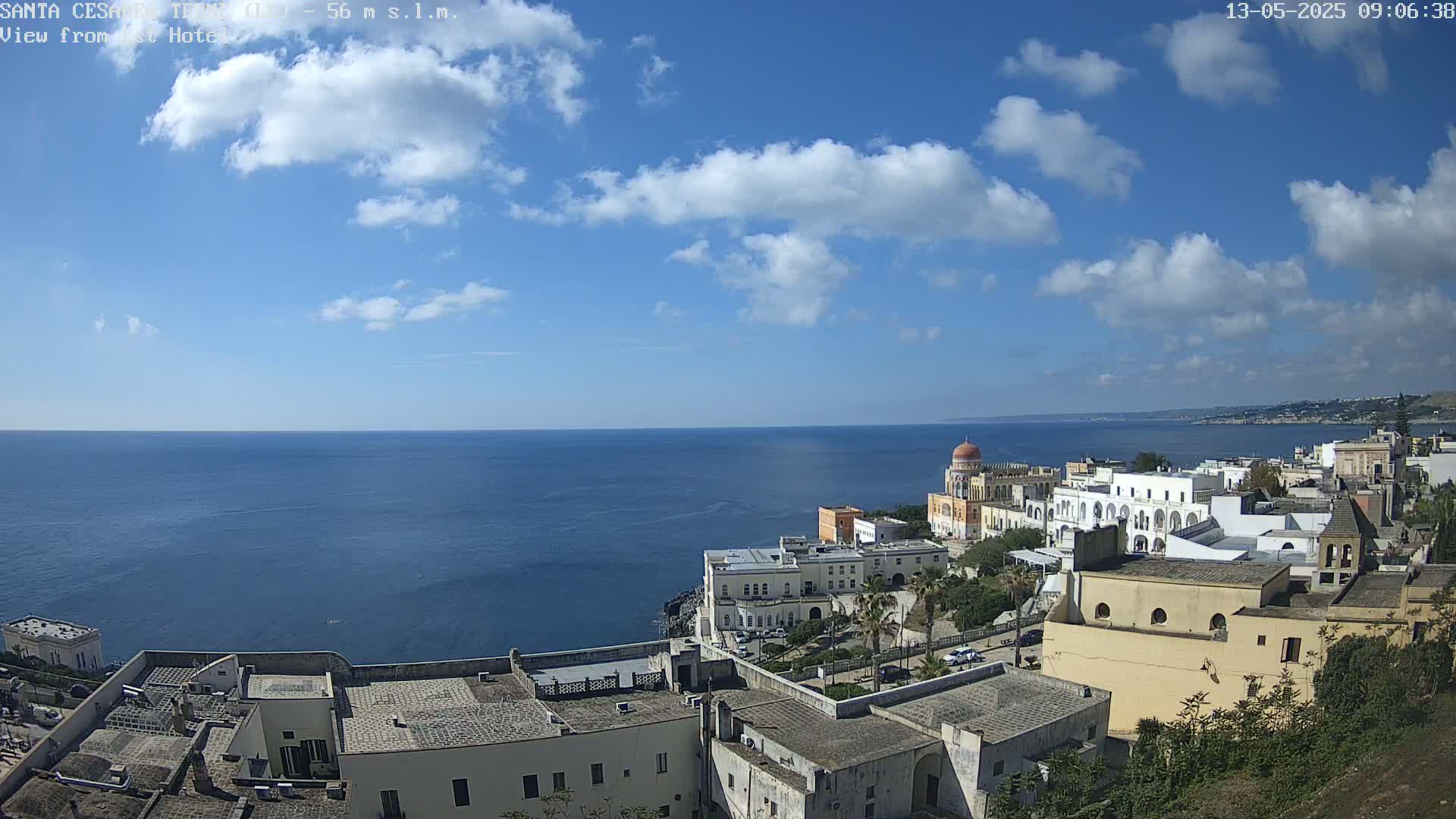 A coastal town with white buildings and a dome-topped structure overlooks a calm blue sea under a mostly sunny sky with scattered cumulus clouds.