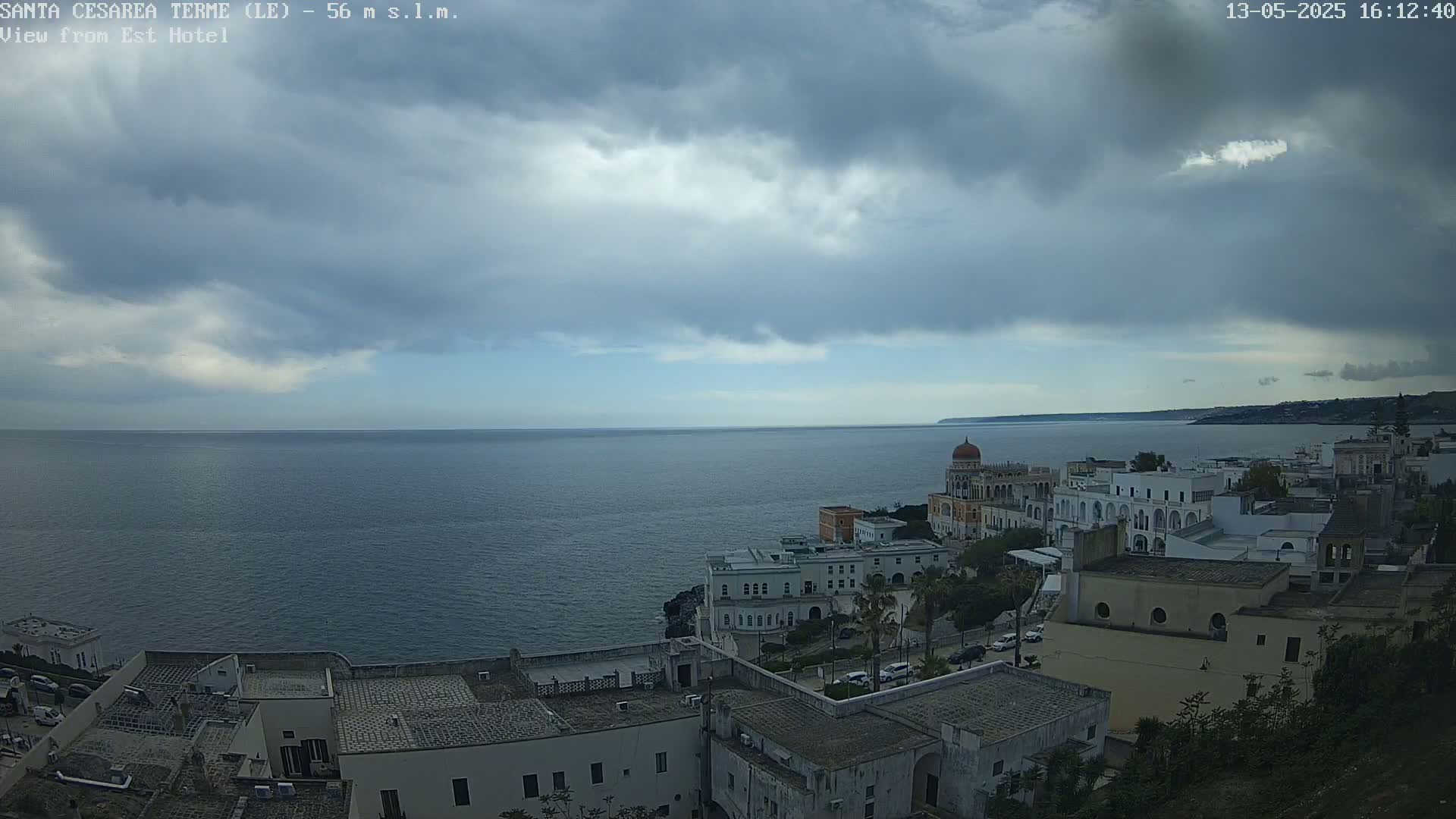 A coastal town with white buildings and a domed structure overlooks a calm sea under a cloudy sky.