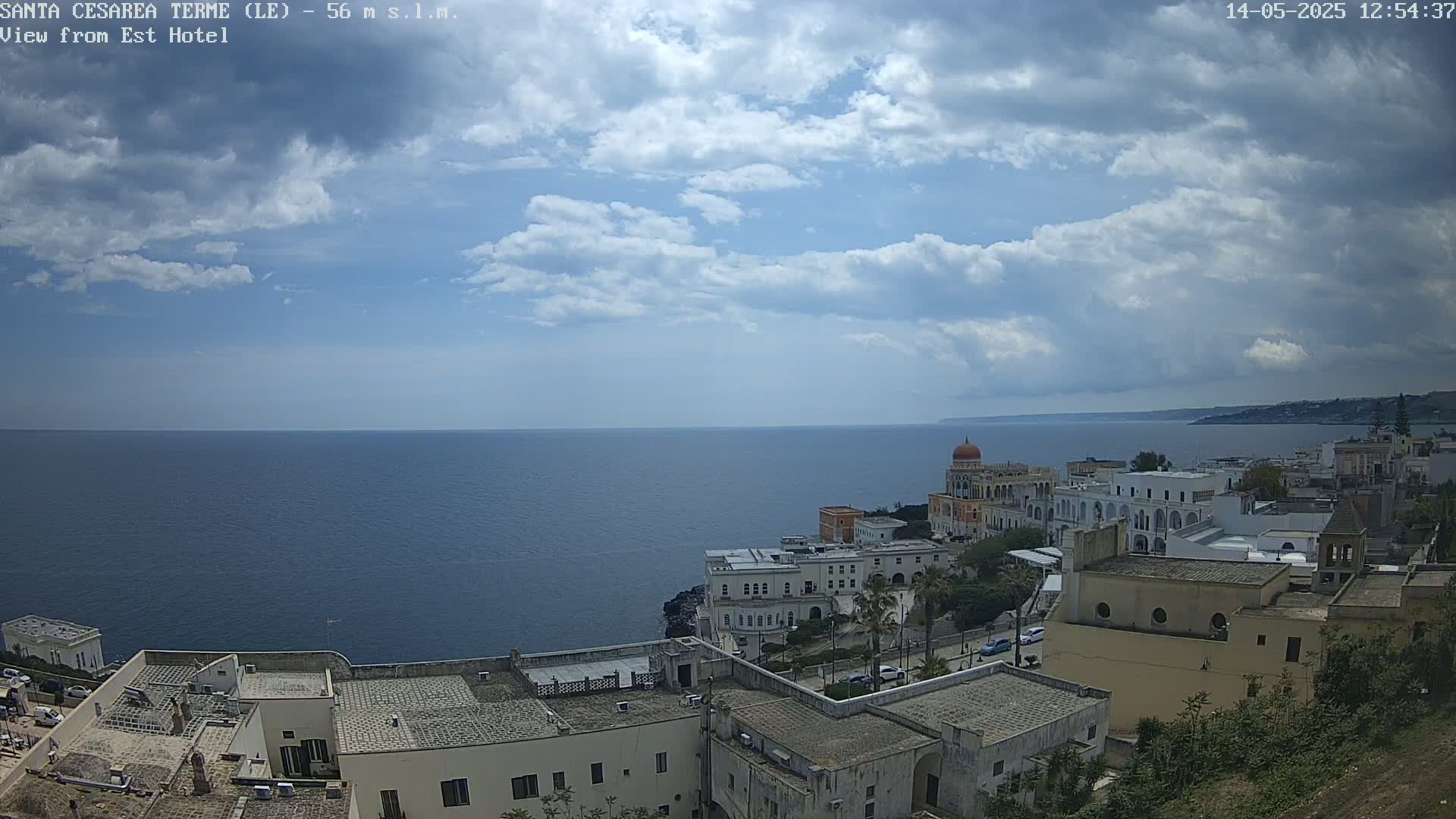 A coastal town with white buildings overlooks a calm blue sea under a partly cloudy sky.