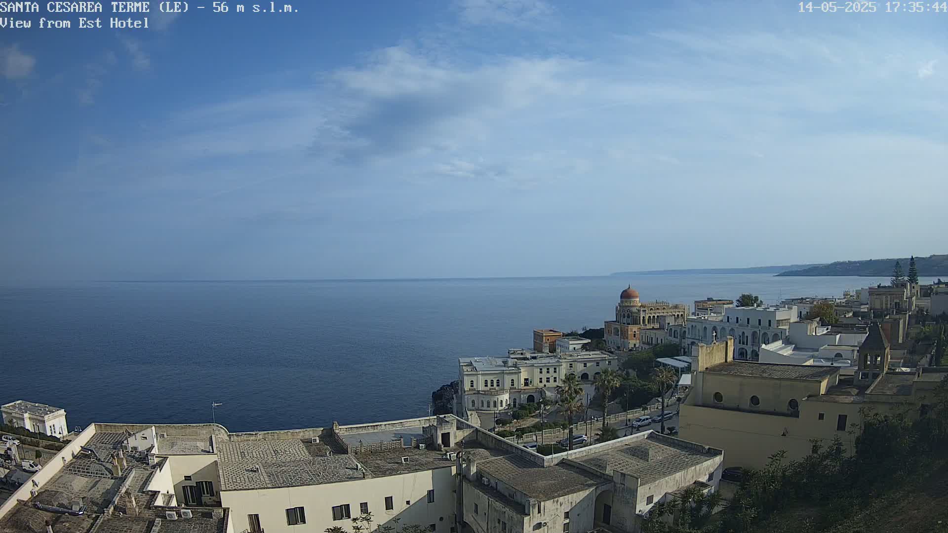 A coastal town with white buildings and a domed structure overlooks a calm, blue sea under a mostly clear sky with a few scattered clouds.
