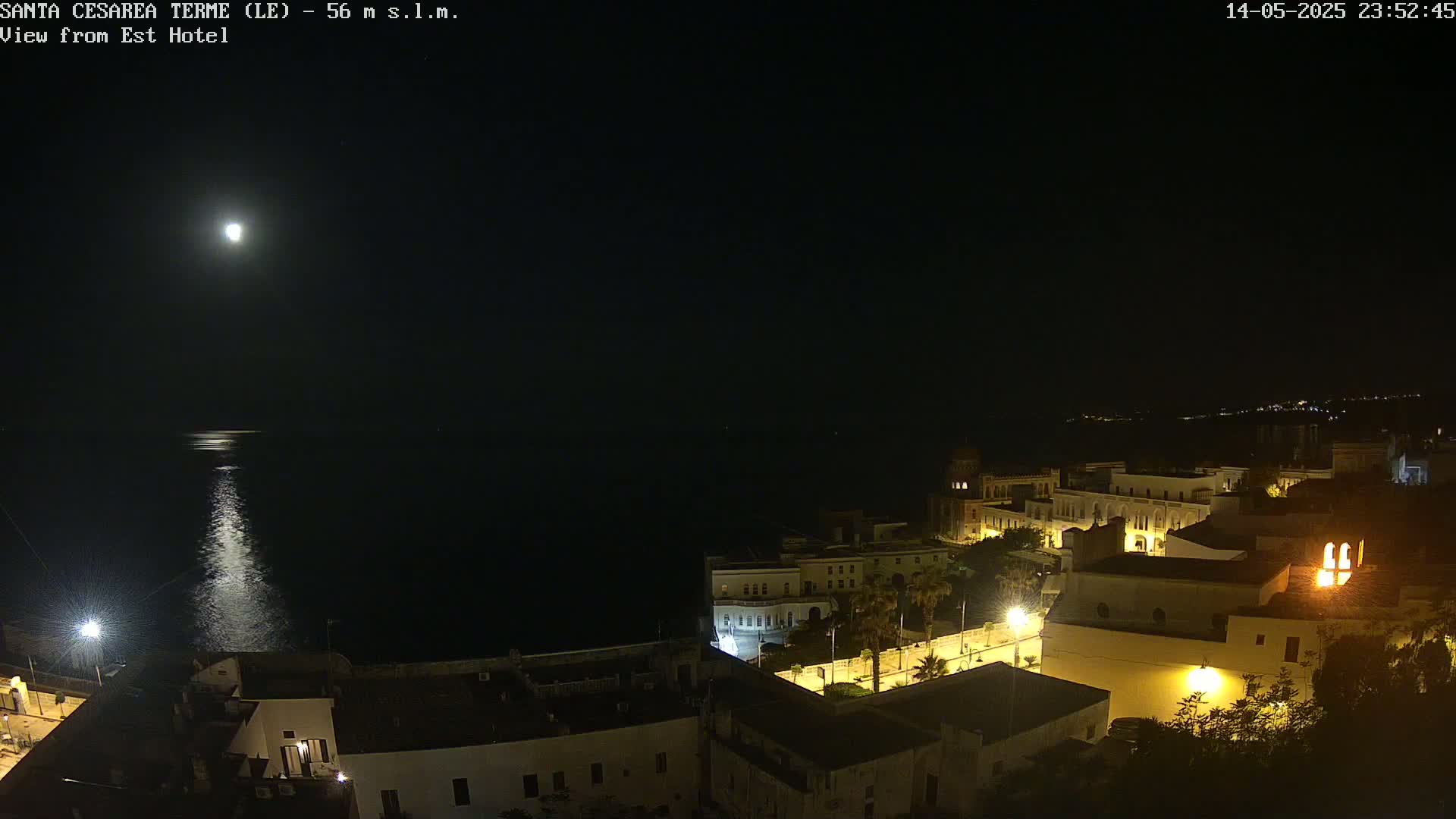 A brightly lit coastal town at night is seen under a full moon, its lights reflecting on the dark water below.