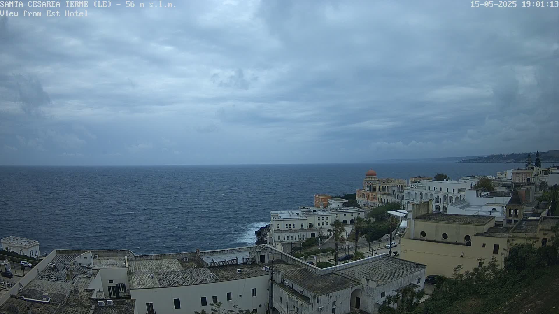 A coastal town with white buildings and a calm, dark-blue sea under a mostly cloudy sky.
