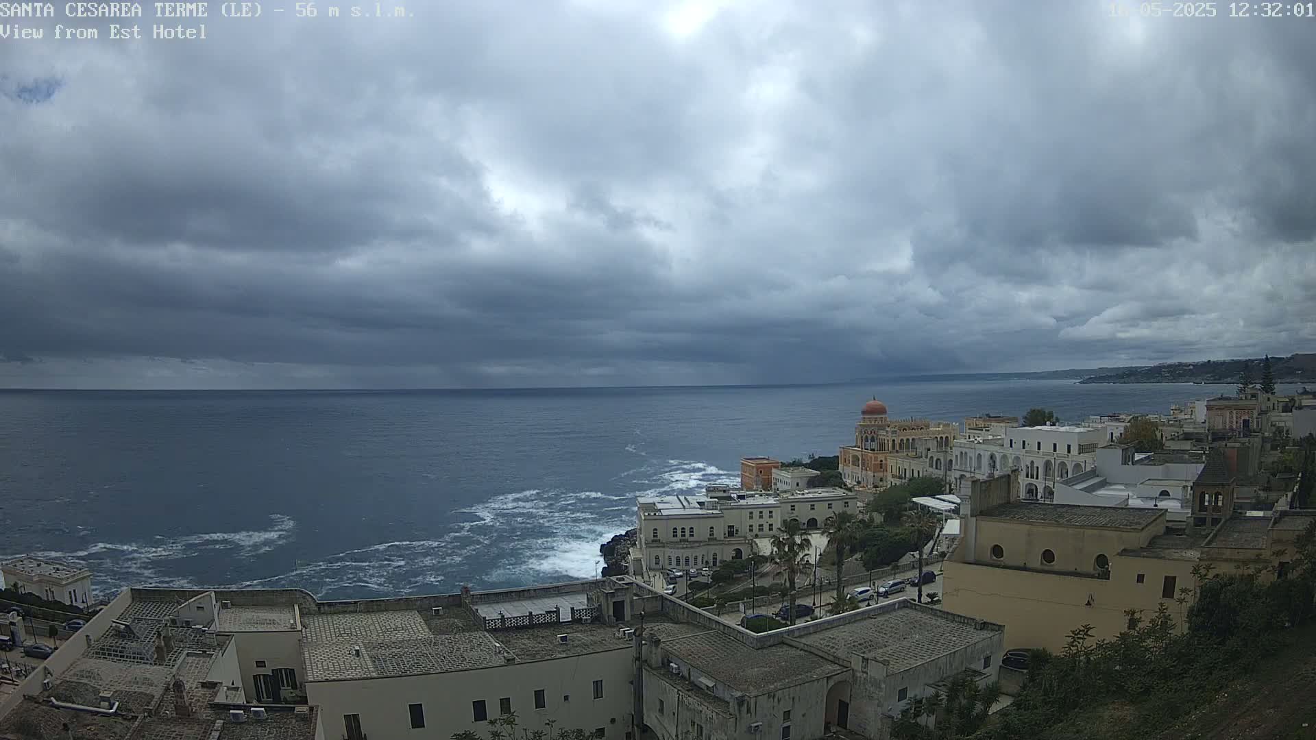 A coastal town with white buildings sits on a cliff overlooking a dark blue sea under a cloudy sky.