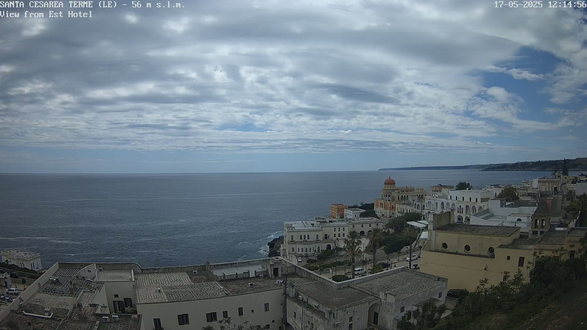 A coastal town with white buildings, a prominent domed structure, and a calm sea under a partly cloudy sky.