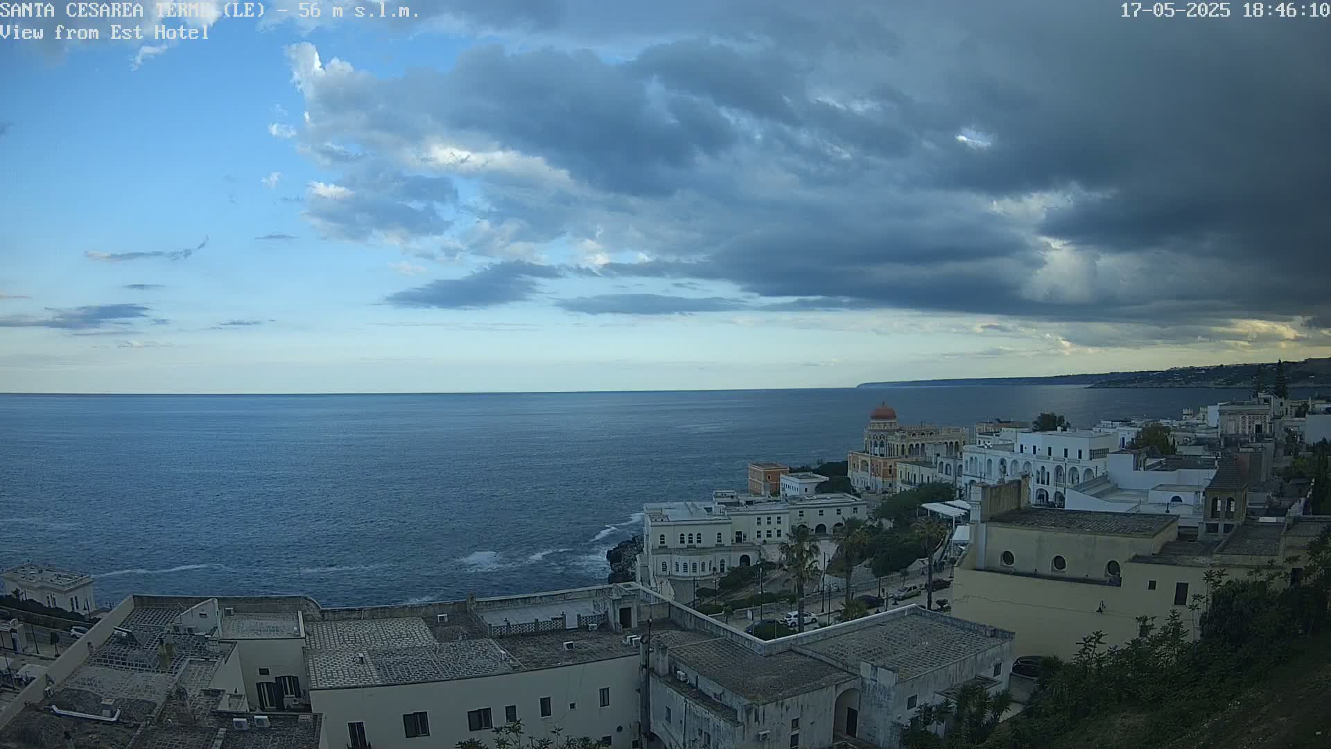 A coastal town with white buildings sits on a cliff overlooking a calm, dark-blue sea under a partly cloudy sky.